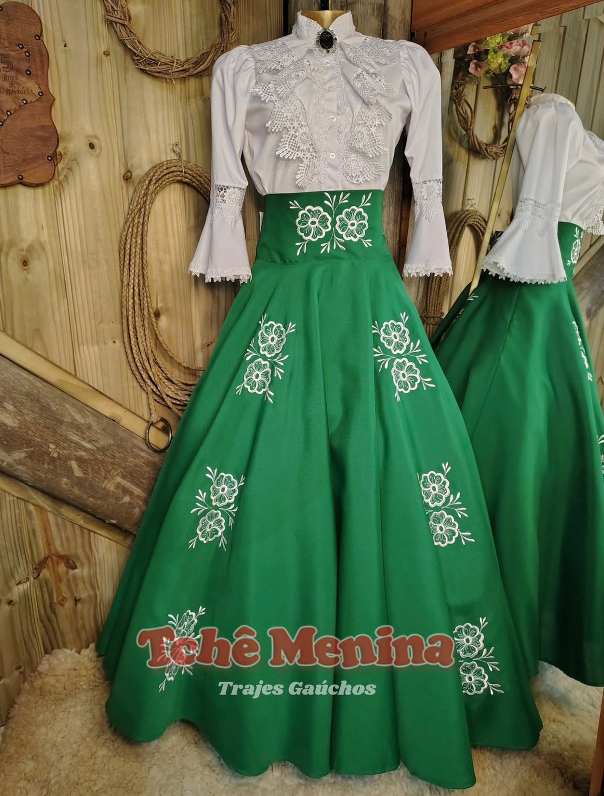 Prenda com vestido de prenda rosa e marinho bordado em cenário de CTG, com roda de carro de boi e bandeira do Rio Grande do Sul